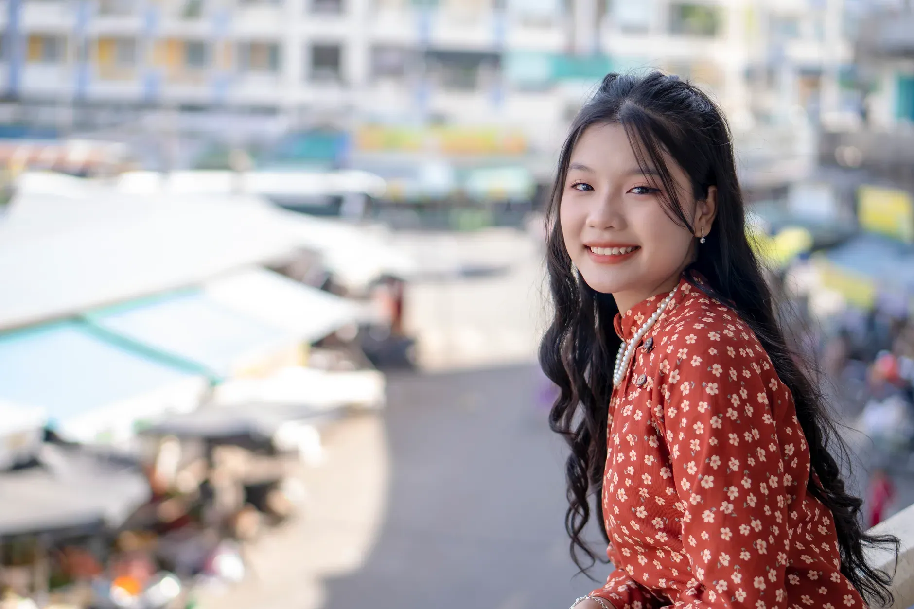 A smiling woman in a floral dress poses in an urban setting, exuding warmth and charm.